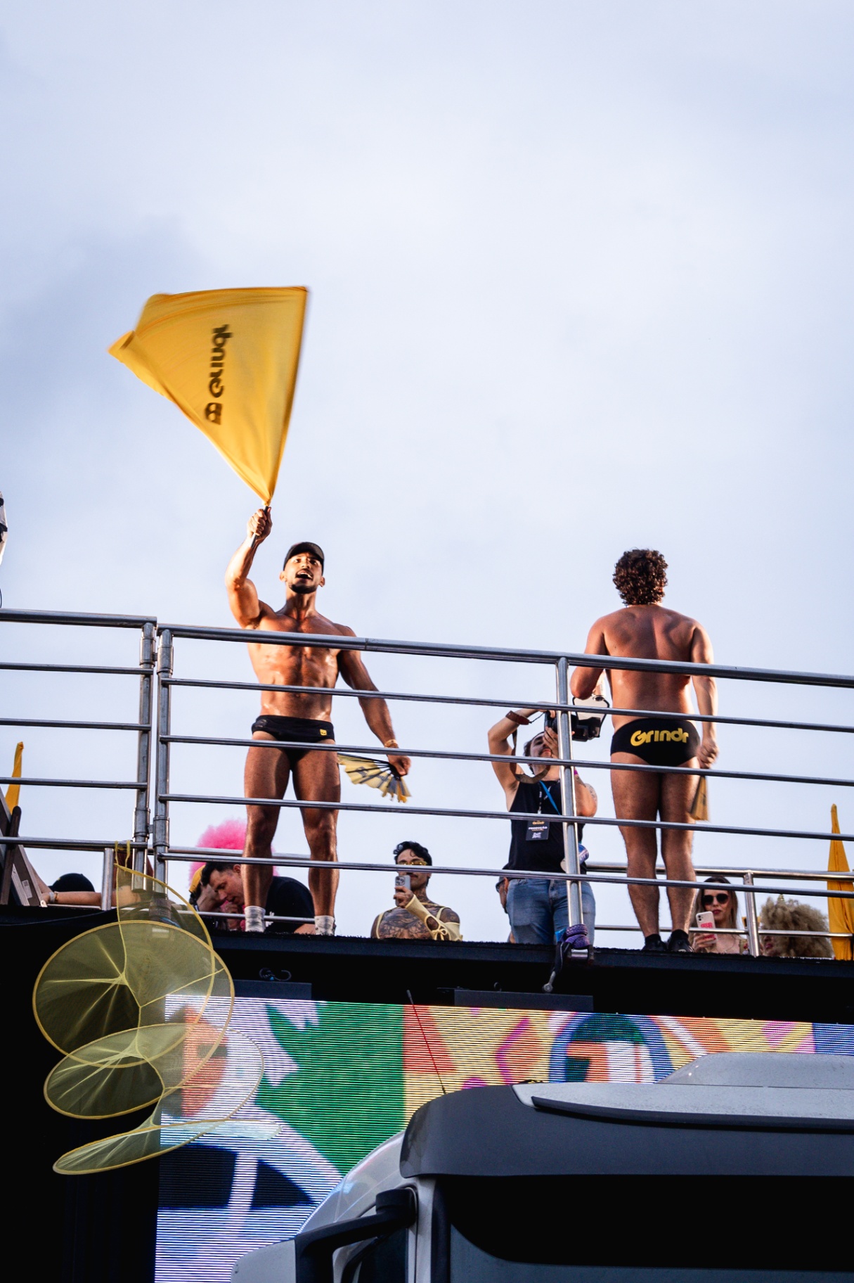 Performers no topo de trio elétrico com bandeira amarela sobre painel de LED colorido